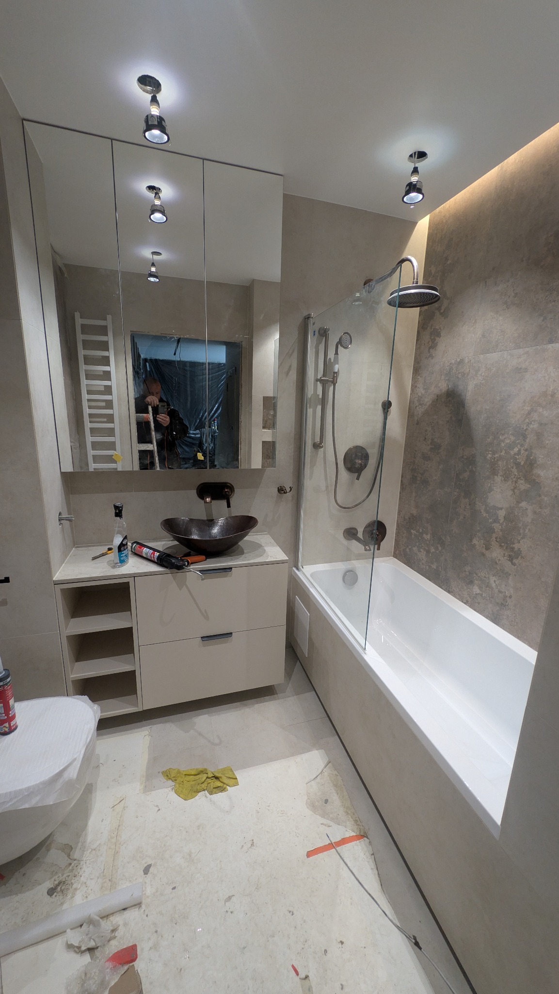 Wnętrze łazienki w trakcie prac wykończeniowych. Widoczna wanna, umywalka z szafką, lustro i częściowo zakryta toaleta. Na podłodze widoczne ślady po remoncie.