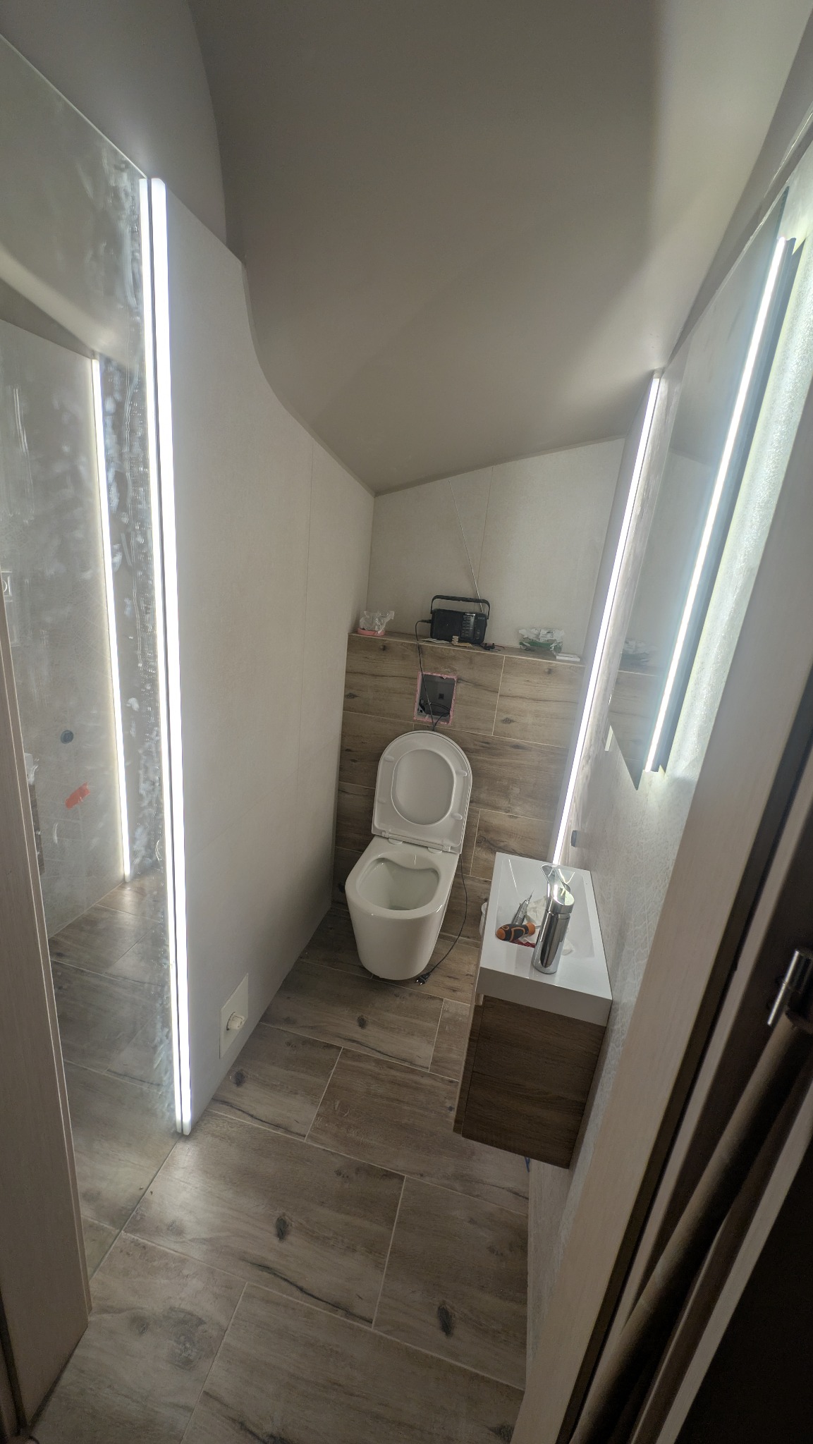 Nowoczesna łazienka z podświetlanymi lustrami, drewnopodobnymi płytkami i wiszącą toaletą. Widoczne narzędzia na umywalce sugerują etap wykończeniowy.