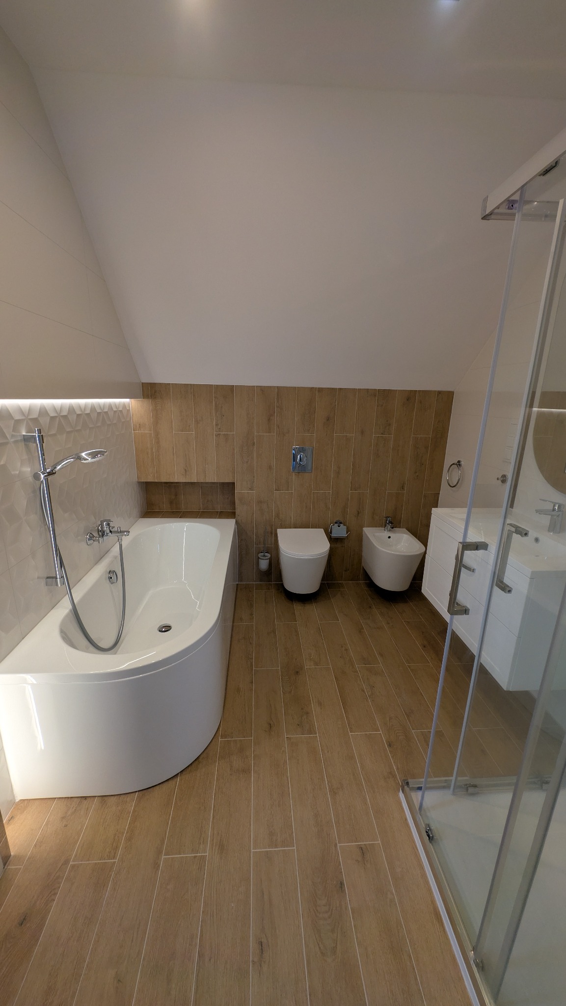 Jasna łazienka na poddaszu z drewnopodobnymi płytkami, białą armaturą i kabiną prysznicową ze szkła. Uwagę przykuwa nietypowy kształt wanny.