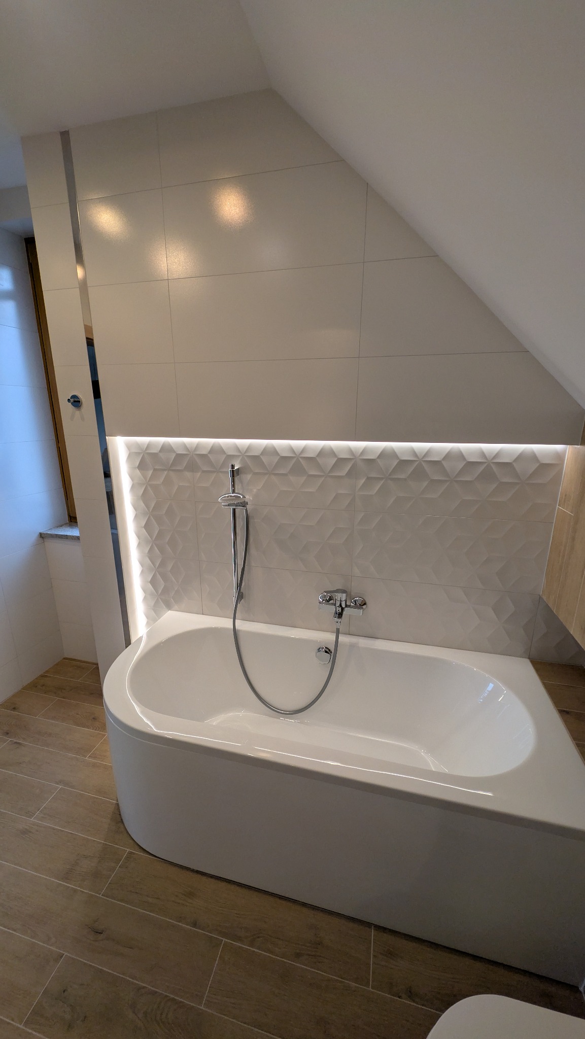 Nowoczesna łazienka na poddaszu z białą wanną, mozaiką 3D i subtelnym oświetleniem LED. Uwagę przykuwają drewnopodobne płytki podłogowe i okno w skosie.