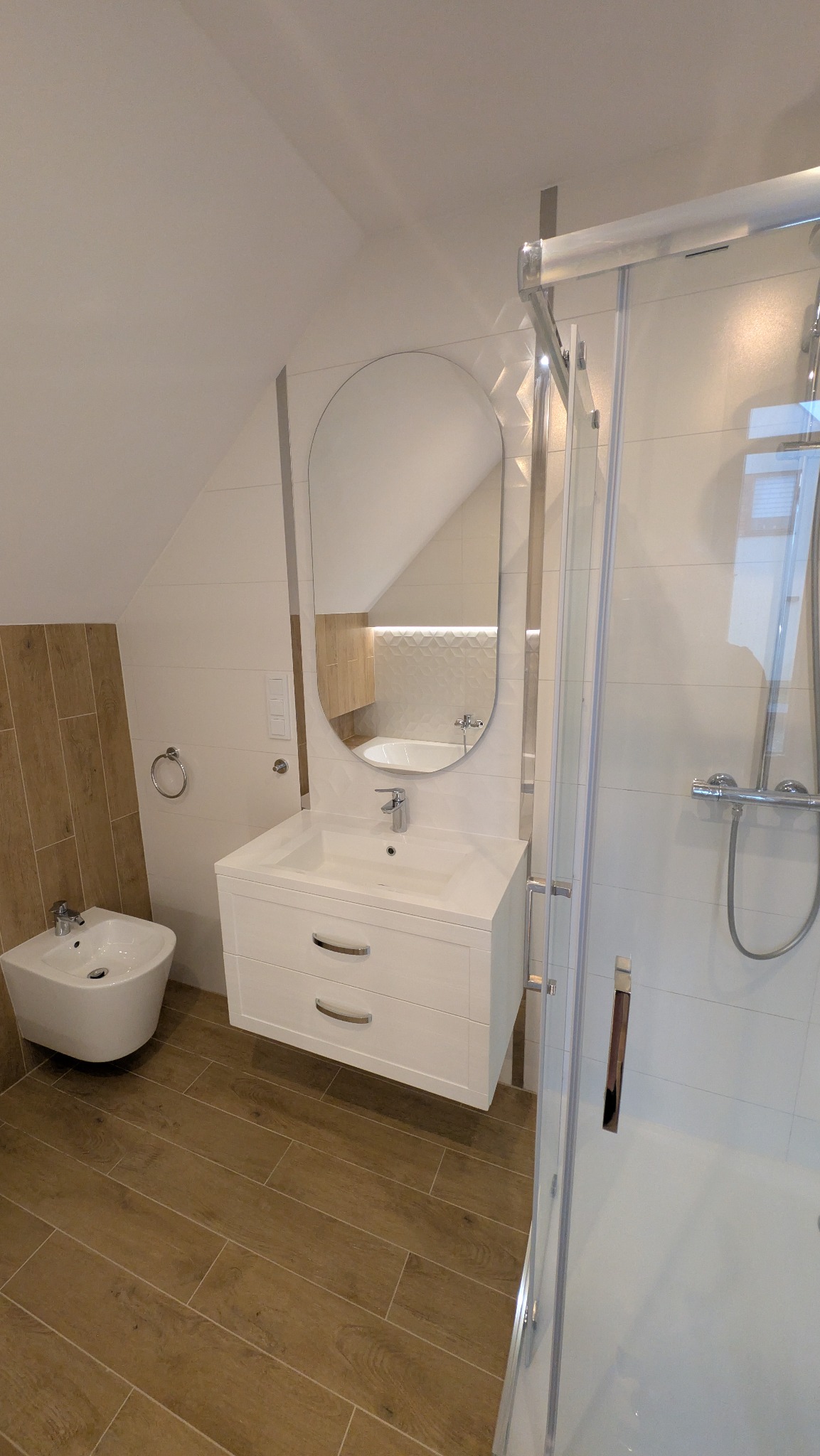 Nowoczesna łazienka na poddaszu z drewnopodobną podłogą, białą szafką z umywalką i owalnym lustrem. Kabina prysznicowa z chromowanymi elementami.