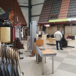 Ekspozycja materiałów budowlanych wewnątrz sklepu: próbki dachówek na miniaturowych konstrukcjach dachowych, panele podłogowe i farby w tle punktu obsługi klienta.