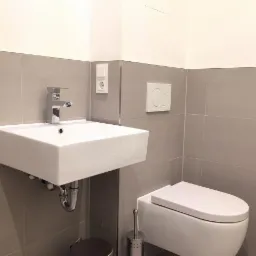 Nowoczesna, minimalistyczna łazienka z umywalką nablatową, chromowaną baterią, szarymi płytkami i podwieszaną toaletą. Widoczny kosz na śmieci i szczotka do WC.