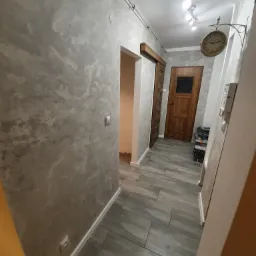 Korytarz po remoncie: ściana z dekoracyjną szarą gładzią, drewniana podłoga, widoczny zegar na ścianie i narzędzia w tle.