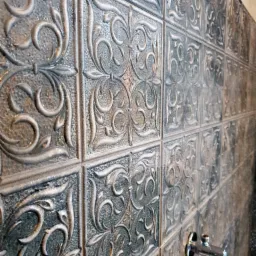 Ściana wyłożona dekoracyjnymi, kwadratowymi płytkami ceramicznymi w odcieniach szarości, z ornamentem roślinnym i widocznymi zaworami wodnymi.