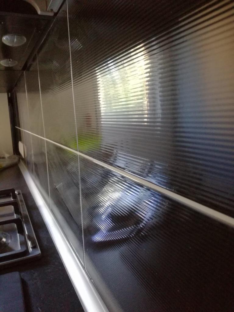 Błyszcząca, czarna ściana nad blatem kuchennym wyłożona płytkami z wyraźną, poziomą fakturą, odbijająca światło okna i lampy, z widoczną częścią kuchenki gazowej.