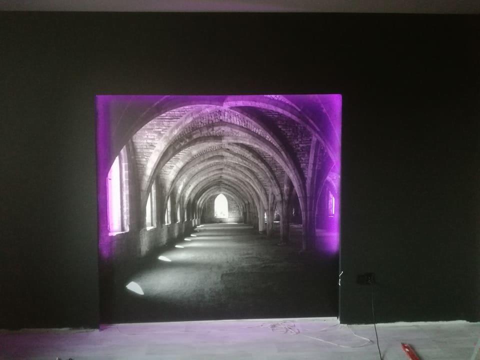 Podświetlony na fioletowo fototapeta z motywem korytarza ze sklepieniami łukowymi na ciemnej ścianie, widoczne białe kable i fragment podłogi.