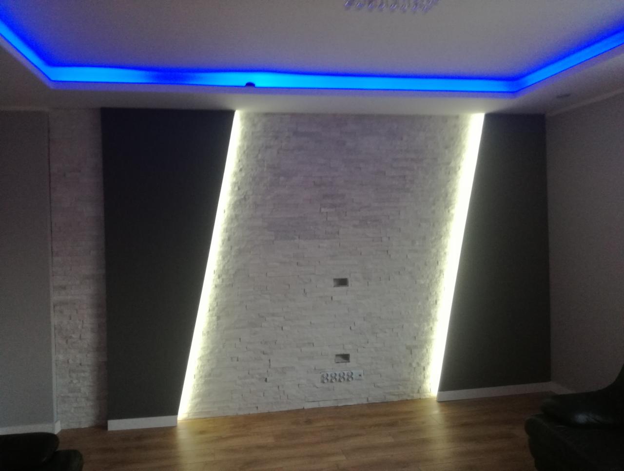 Ściana z białej cegły dekoracyjnej z pionowym podświetleniem LED, czarne panele po bokach, drewniana podłoga i niebieskie oświetlenie sufitowe.