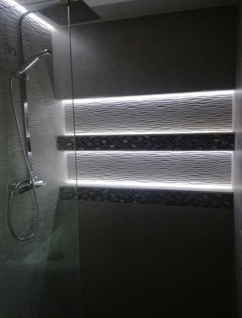 Wykończona łazienka z szarymi płytkami falistymi, podświetleniem LED w niszach i mozaiką, kabina prysznicowa ze szklaną ścianką i deszczownicą.