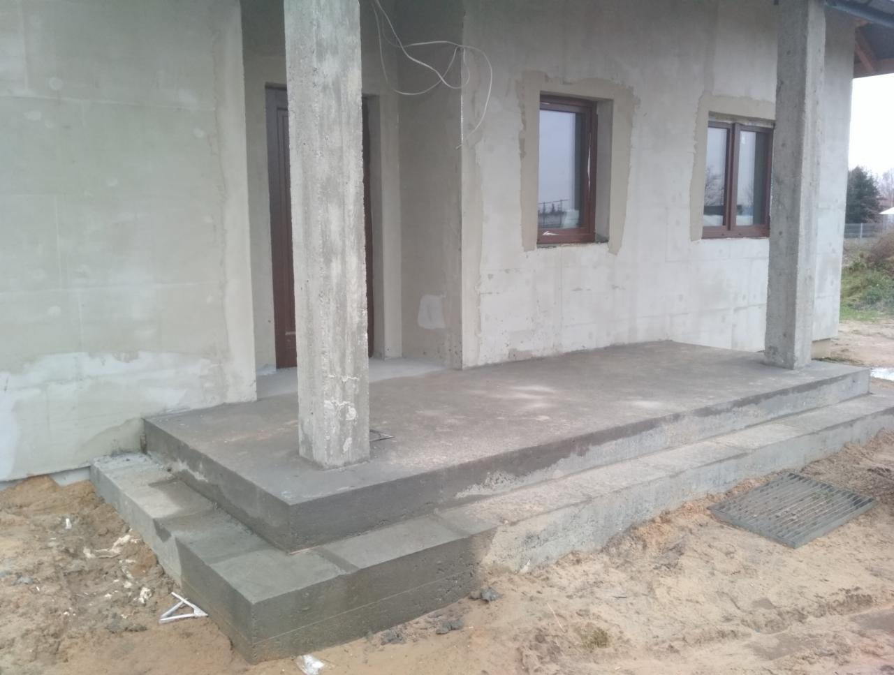 Surowy stan domu jednorodzinnego z widocznym betonowym tarasem, schodami wejściowymi i filarami podtrzymującymi zadaszenie, okna w brązowych ramach, otoczenie w stanie budowy.