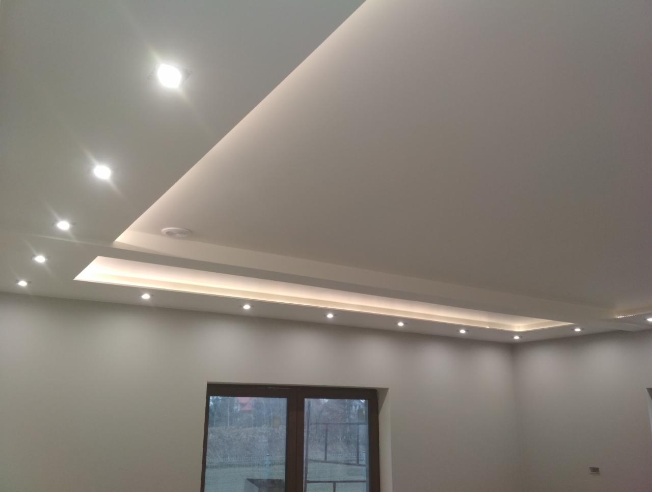 Oświetlenie sufitowe z wbudowanymi punktami świetlnymi i podświetleniem LED, widok na szare ściany i okno.