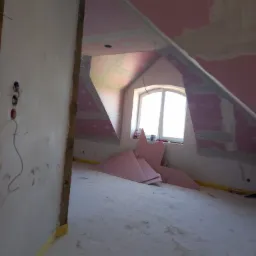 Poddasze w trakcie remontu: widoczne płyty kartonowo-gipsowe na skośnych ścianach, fragment okna dachowego i rozłożone płyty izolacyjne na podłodze, przygotowane do dalszych prac wykończeniowych.