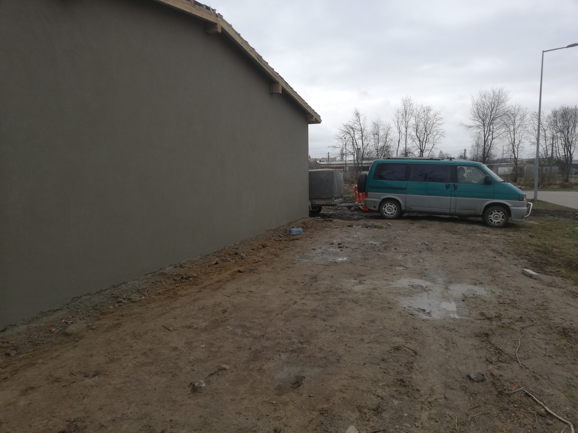 Świeżo otynkowana ściana budynku z widocznymi drewnianymi elementami dachu, obok zaparkowany zielono-szary bus z przyczepką na błotnistym terenie.