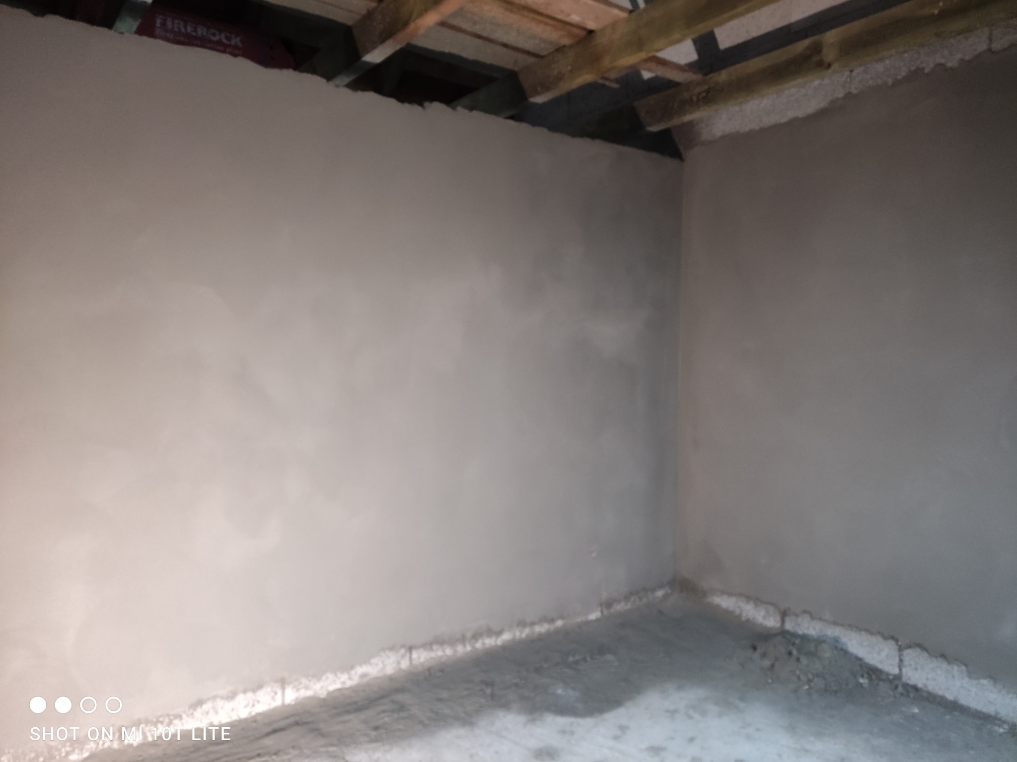 Wnętrze pomieszczenia po nałożeniu świeżego, jasnoszarego tynku na ściany, widoczne surowe drewniane elementy konstrukcyjne stropu oraz fragment etykiety FIRE ROCK na górze kadru.