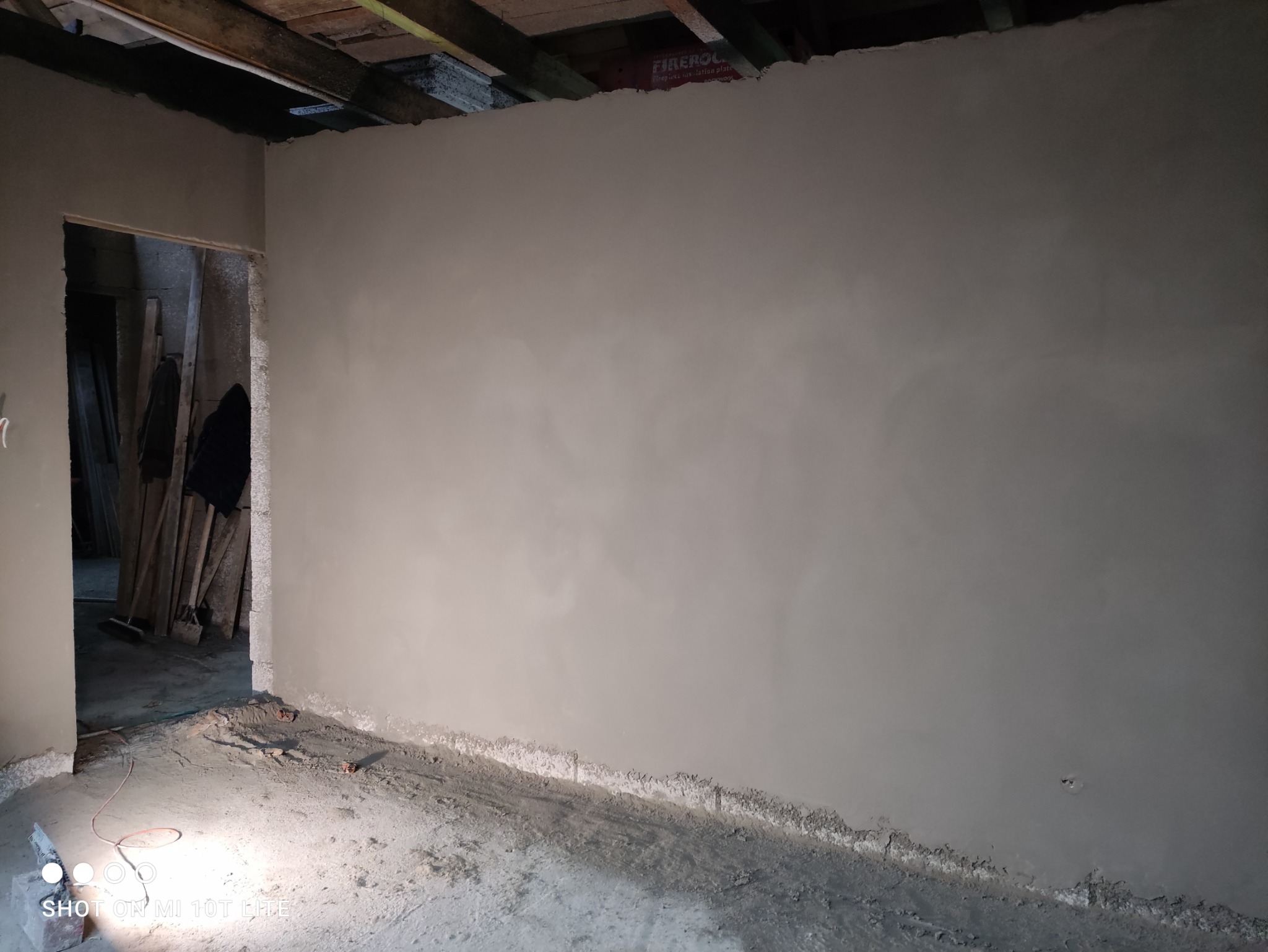 Świeżo otynkowana ściana w surowym wnętrzu, widoczne drewniane elementy konstrukcyjne stropu i otwór drzwiowy z widokiem na składowane materiały budowlane.