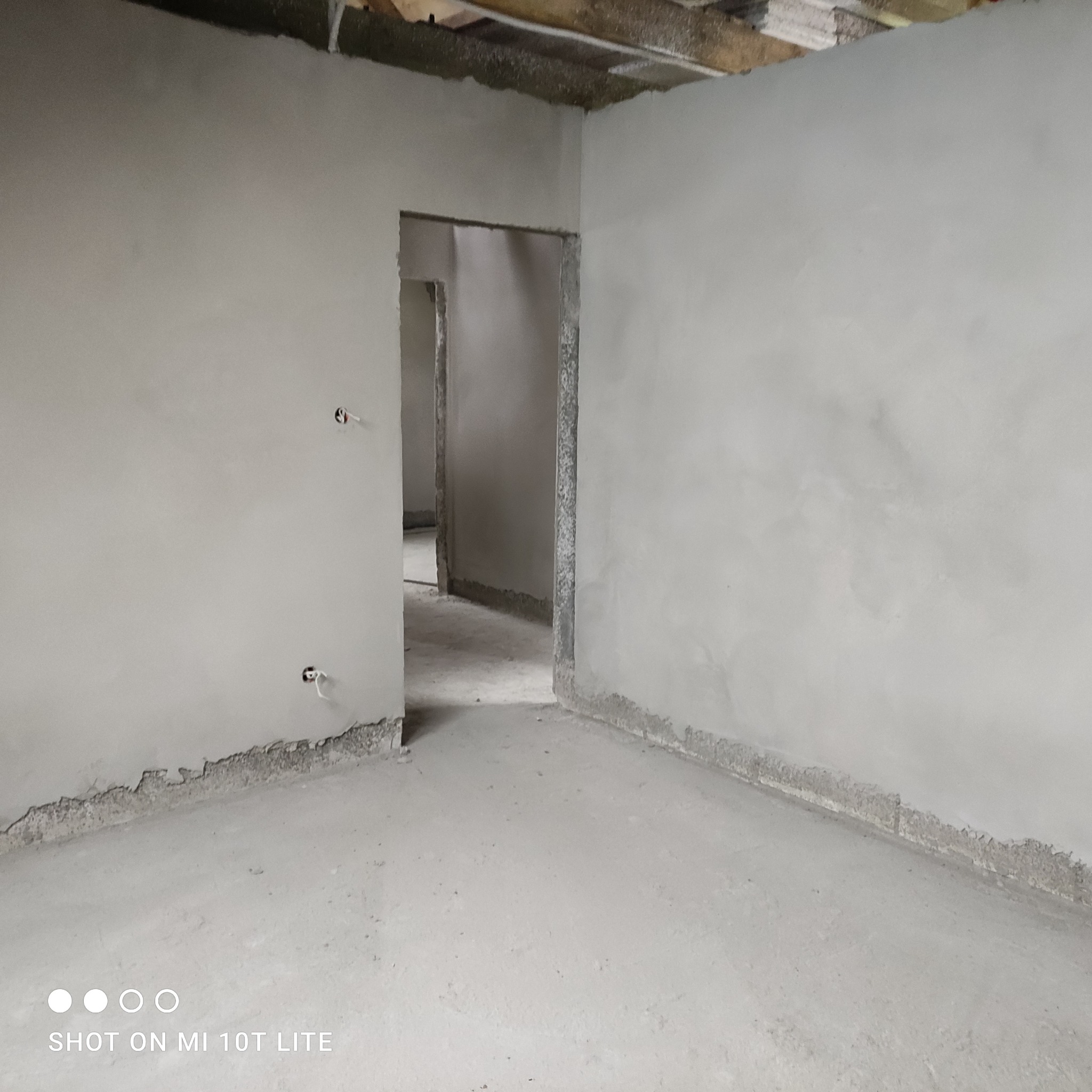 Wnętrze pomieszczenia po nałożeniu szarego tynku, widoczne wystające kable elektryczne ze ściany oraz surowy stan podłogi i fragmentu ściany przy podłodze.