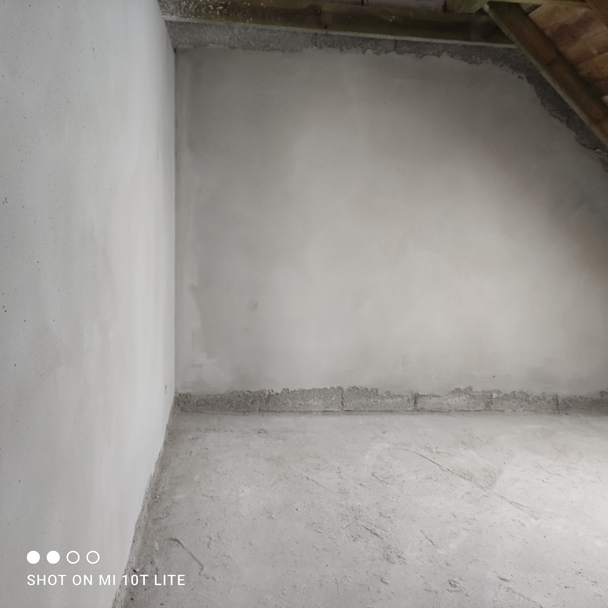 Surowe wnętrze poddasza po tynkowaniu maszynowym, widoczne świeżo otynkowane ściany w odcieniach szarości i betonowa podłoga, fragment drewnianej konstrukcji dachu w prawym górnym rogu.