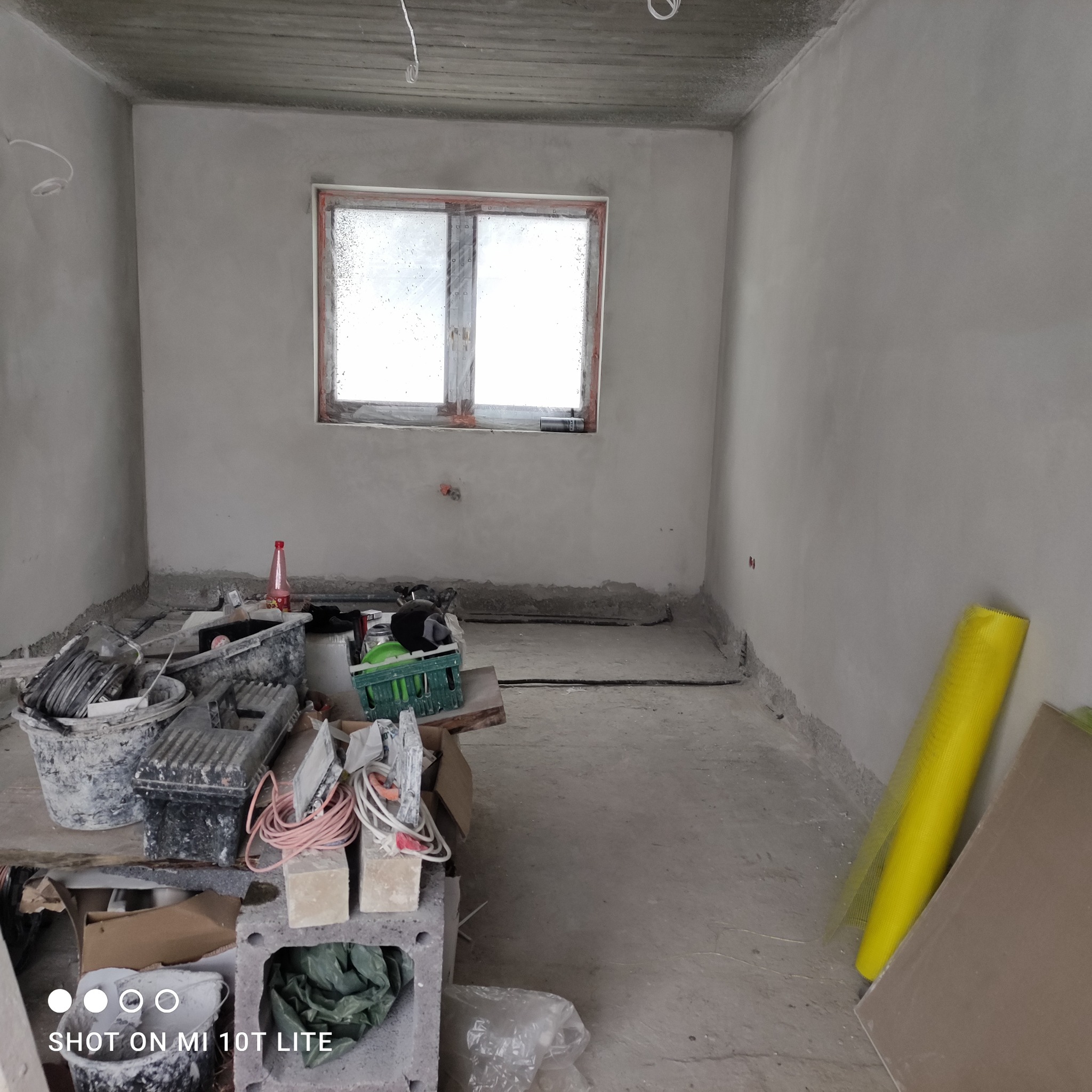 Surowe wnętrze po nałożeniu tynku, widoczne narzędzia i materiały budowlane, okno z widocznymi kroplami wody, ściany pokryte świeżym tynkiem.