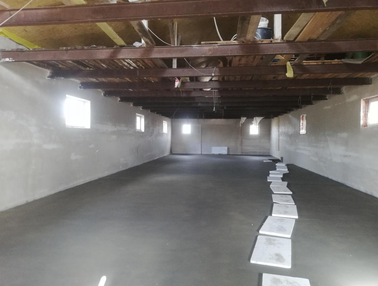 Wnętrze budynku w trakcie prac wykończeniowych: świeżo wylana posadzka betonowa, tynkowane ściany z oknami, surowy drewniany strop z elementami metalowymi, płyty chodnikowe ułożone na posadzce.