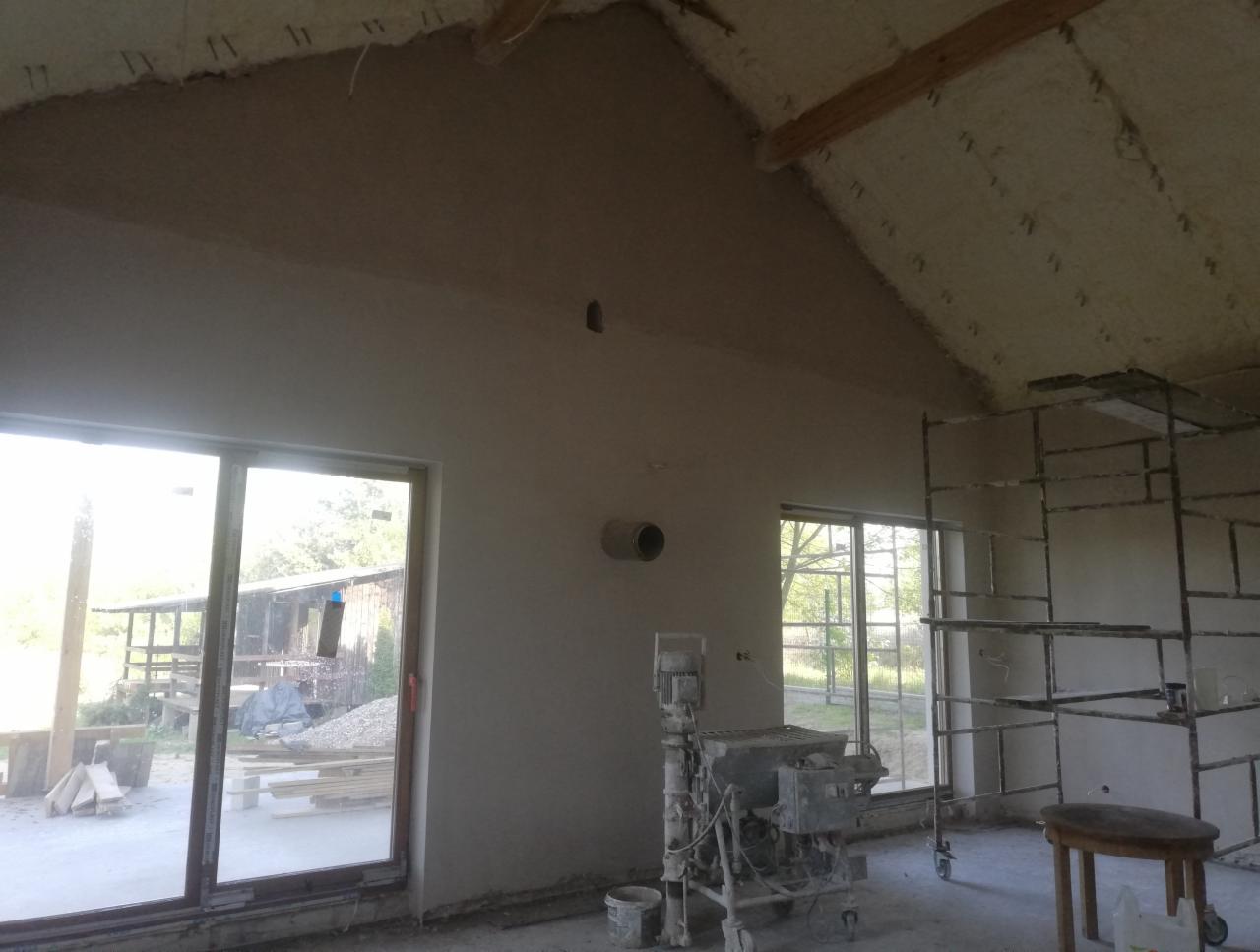 Wnętrze budynku w trakcie prac wykończeniowych, ściany pokryte świeżym tynkiem, widoczna maszyna do tynkowania i rusztowanie, sufit ocieplony pianą, okna z widokiem na zewnątrz.