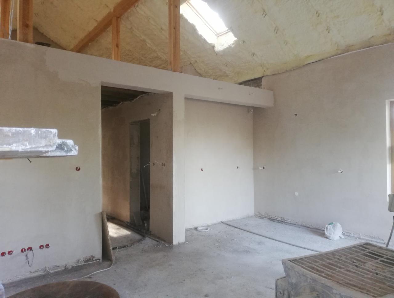 Wnętrze w trakcie tynkowania maszynowego: ściany pokryte świeżym tynkiem, widoczne instalacje elektryczne, pianka izolacyjna na poddaszu, fragment rusztowania i resztki materiałów na podłodze.