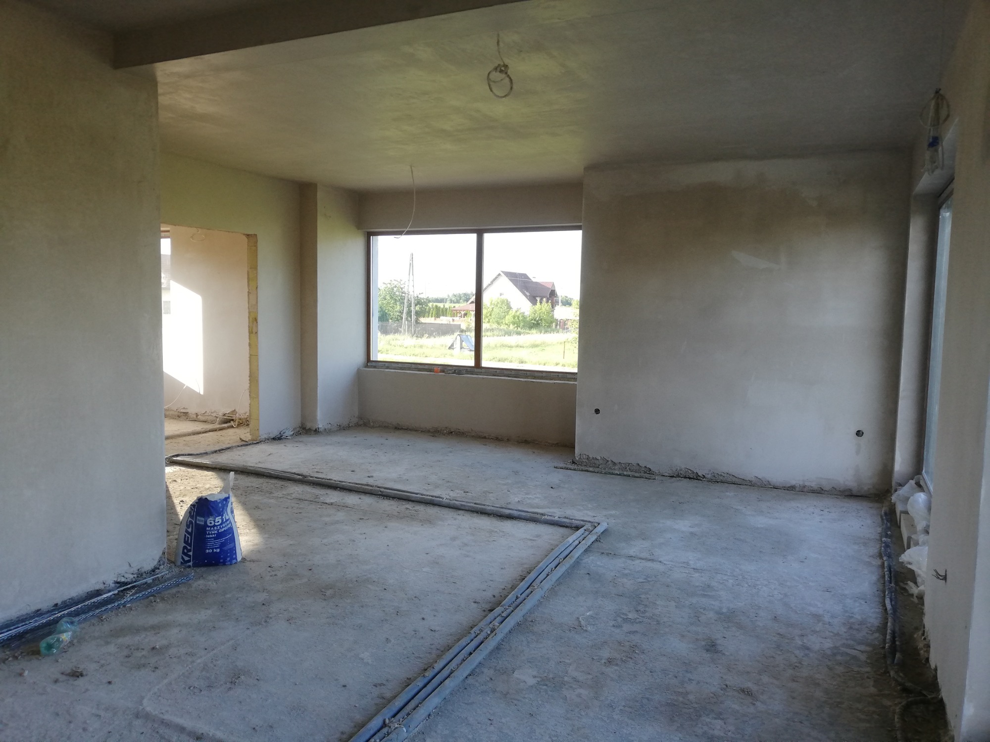 Surowe wnętrze po nałożeniu tynków w Nidzicy, z workiem zaprawy gipsowej Kreisel 651 i rurami instalacyjnymi na podłodze, w tle widok na wiejski krajobraz za oknem.