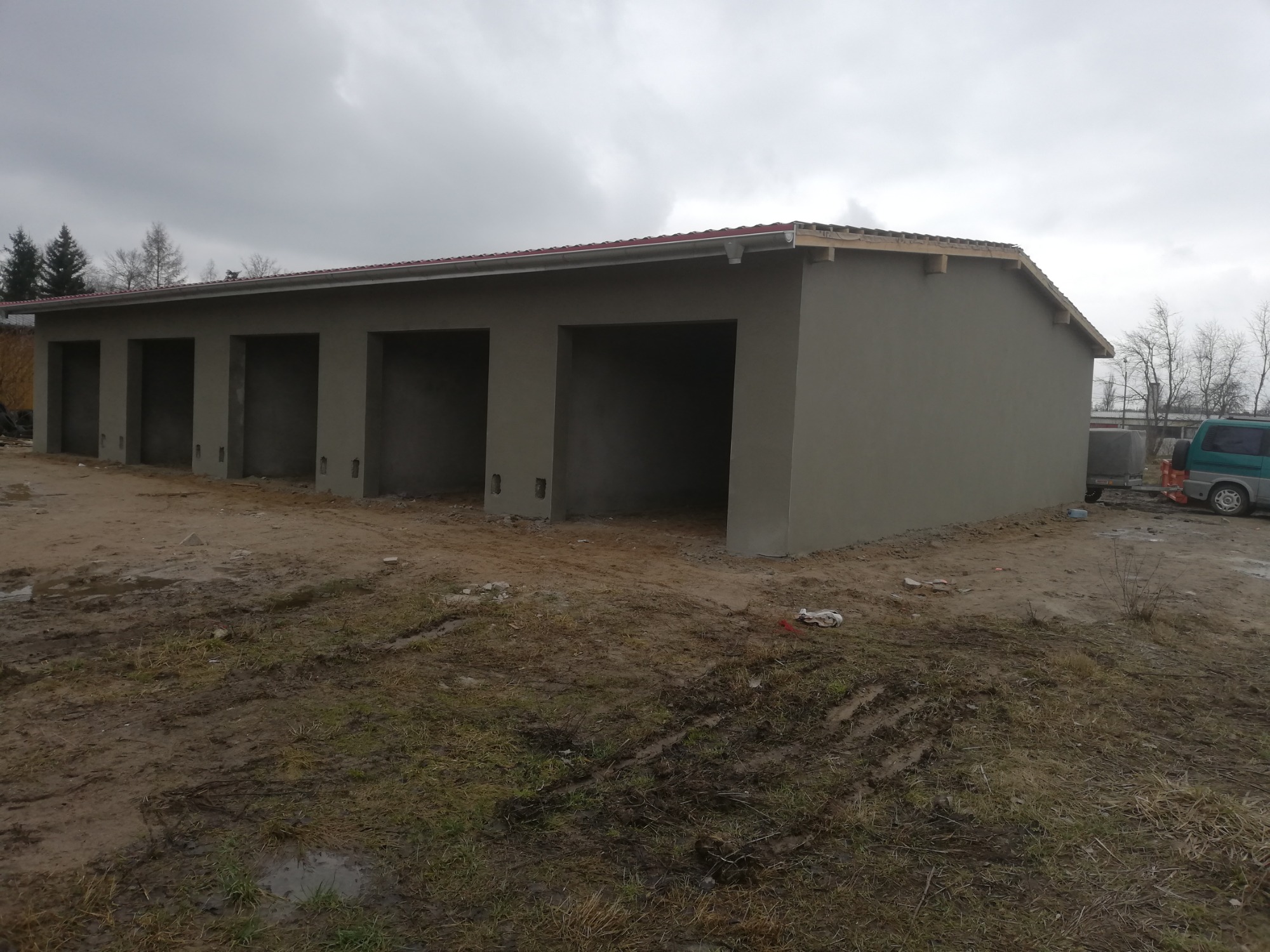 Nowo otynkowany budynek garażowy z czterema stanowiskami, widok zewnętrzny w pochmurny dzień, teren wokół w stanie surowym, widoczne ślady opon.