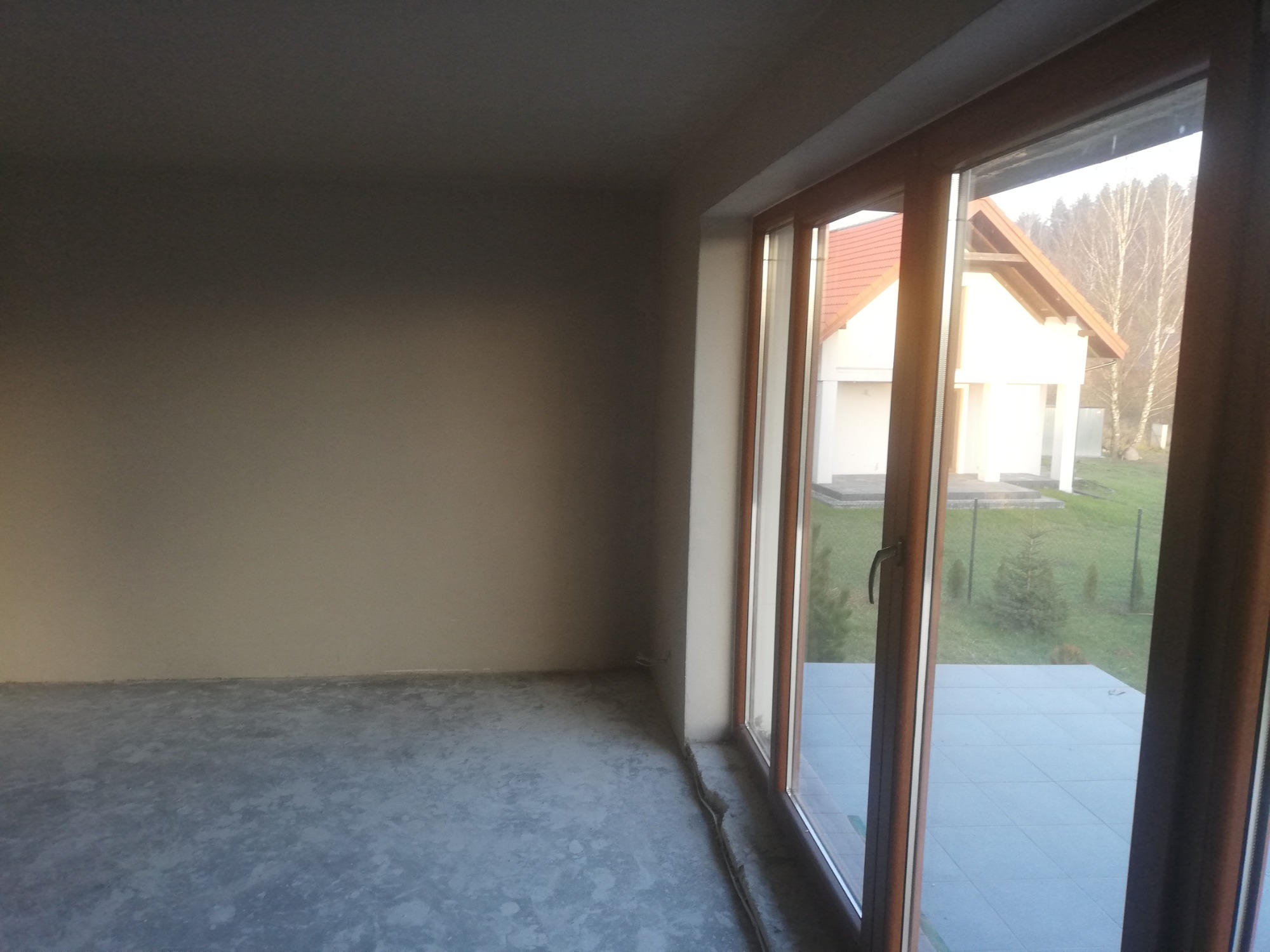 Surowe wnętrze z widokiem na dom przez duże okno balkonowe; ściany przygotowane pod tynkowanie, szara wylewka na podłodze, brak wykończenia.
