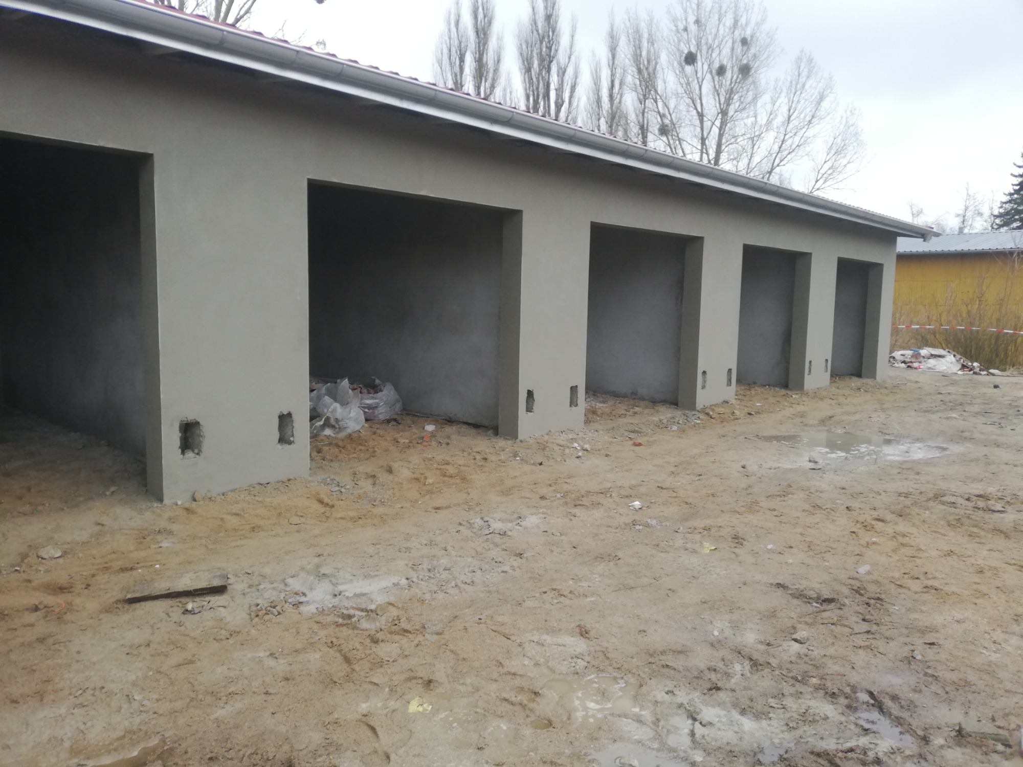 Surowy budynek garażowy z świeżo nałożonymi tynkami zewnętrznymi, widoczne otwory w ścianach i worki z materiałami wewnątrz, teren wokół w stanie surowym.