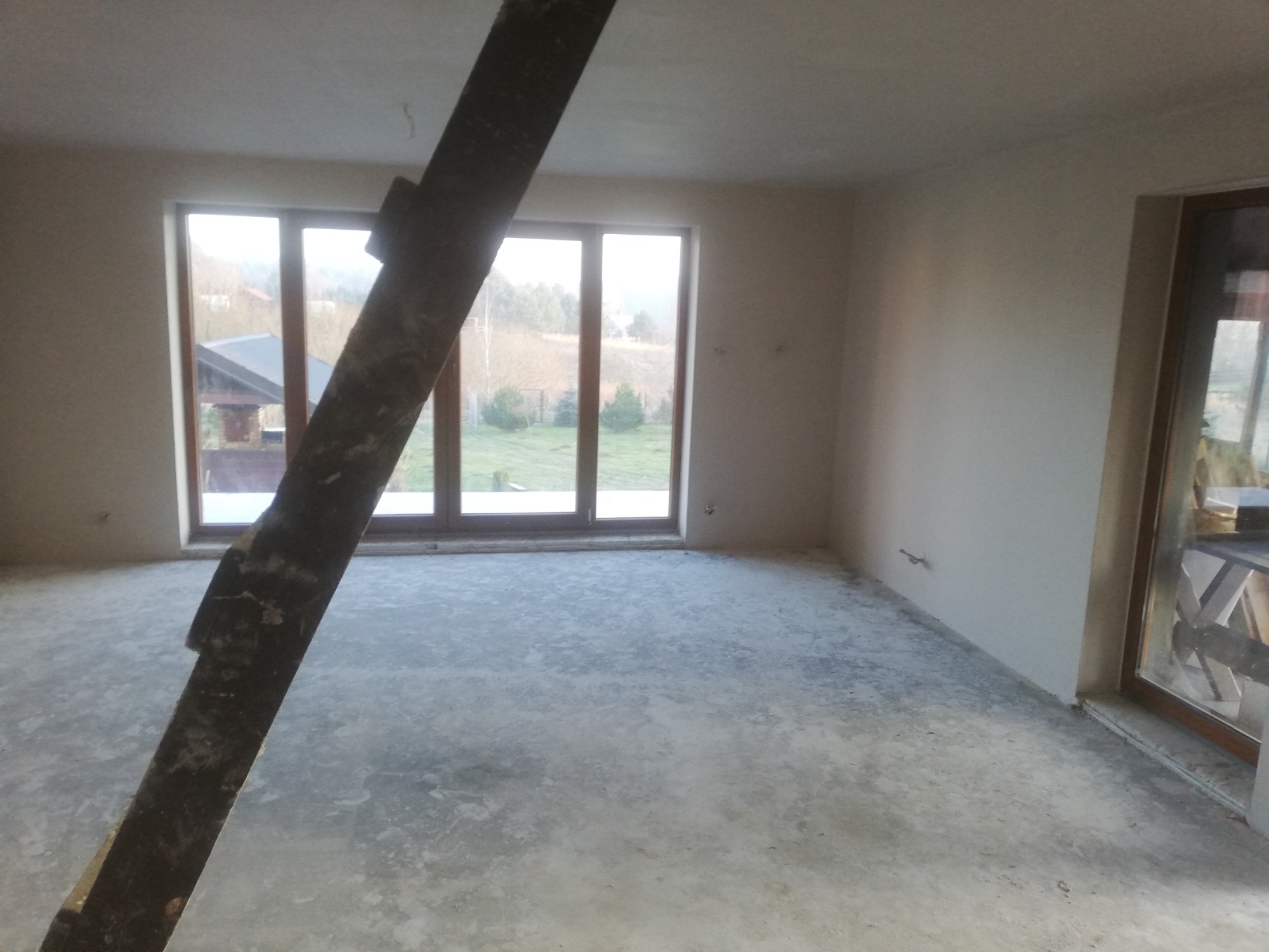 Surowe wnętrze w trakcie remontu: betonowa podłoga, surowe ściany przygotowane do tynkowania, drewniana belka podpierająca strop na pierwszym planie, duże okno tarasowe z widokiem na ogród.