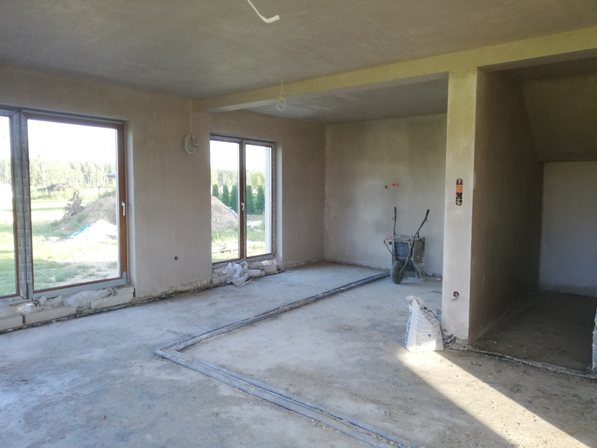 Wnętrze surowego domu po nałożeniu tynków, z widocznymi dwoma oknami, bruzdami w podłodze, taczką i workiem zaprawy.