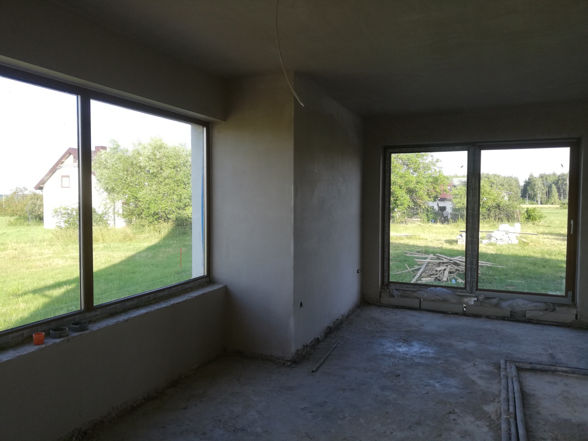 Surowe wnętrze pokoju po nałożeniu tynku maszynowego, widoczne okna z widokiem na zieleń, betonowa podłoga i wystające przewody.