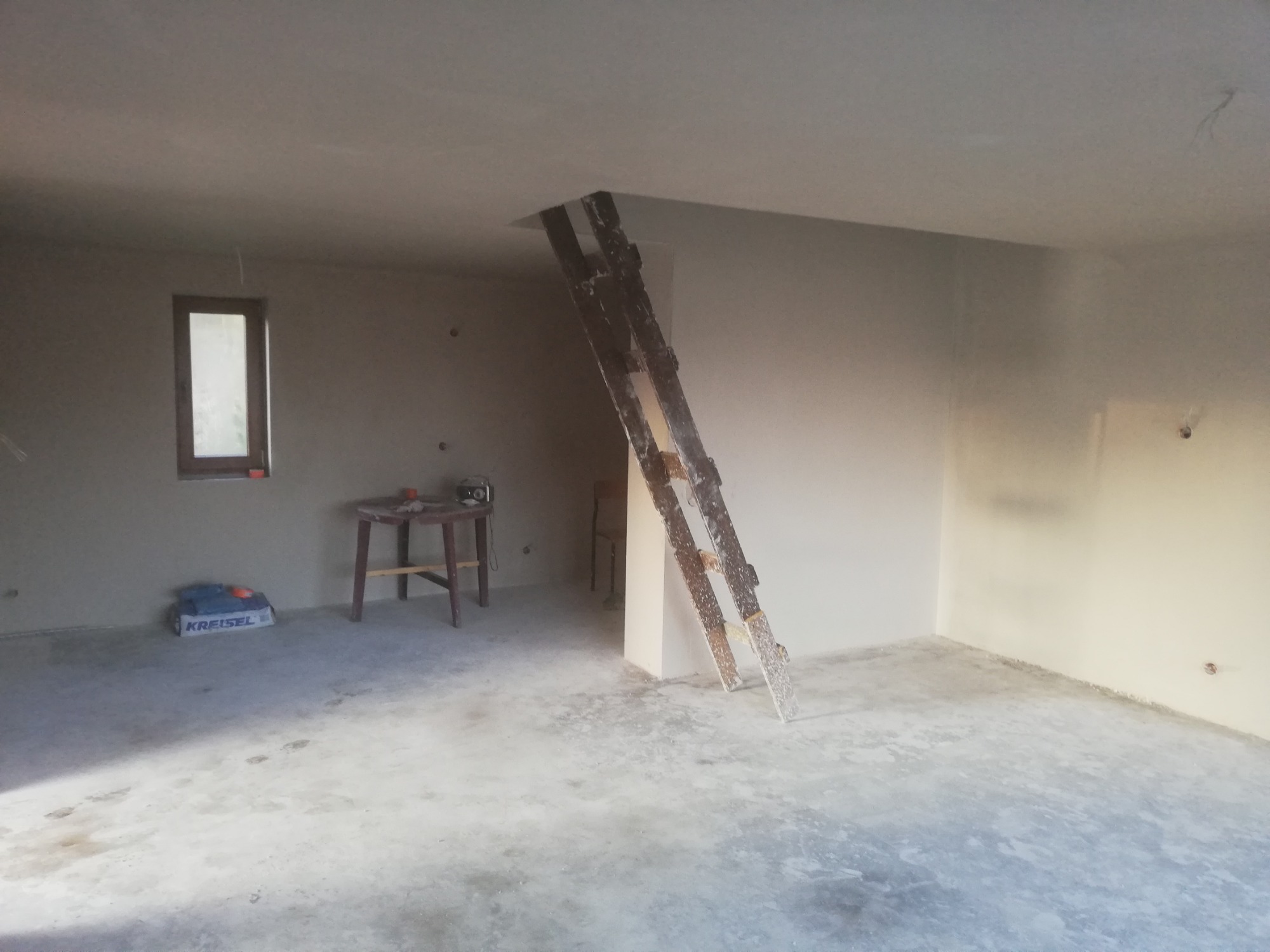 Wnętrze w trakcie prac wykończeniowych: świeżo otynkowane ściany, drewniana drabina oparta o sufit, worek zaprawy Kreisel, mały stolik z narzędziami przy oknie.