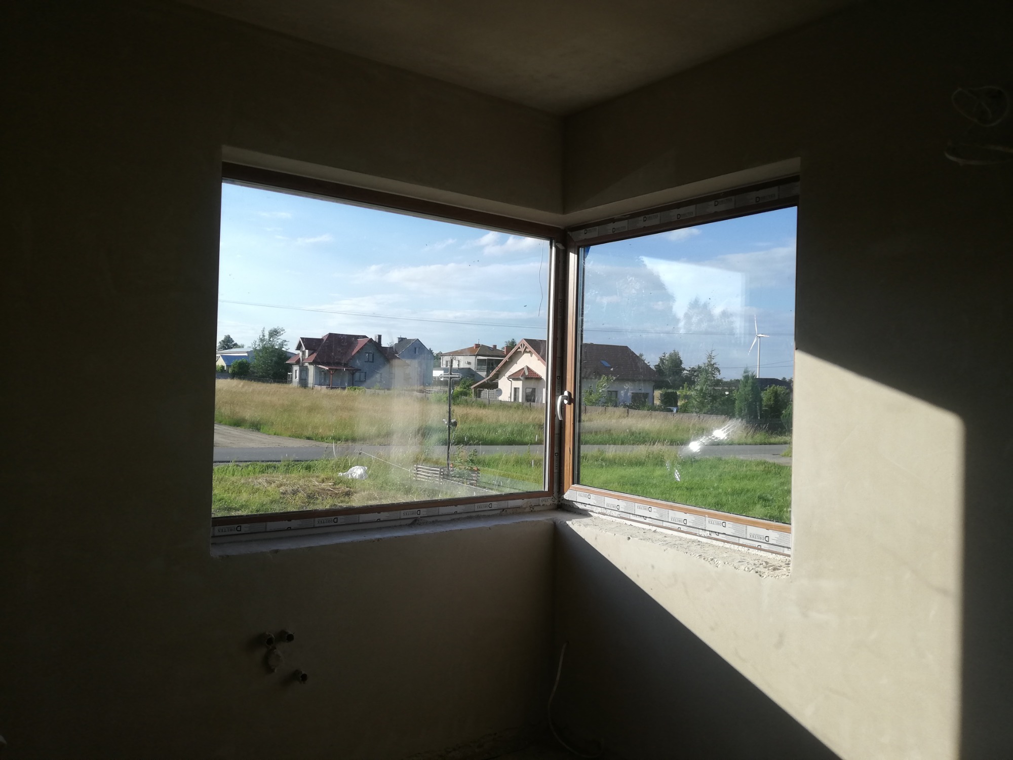 Wnętrze surowego budynku z oknem narożnym, ściany pokryte świeżym tynkiem, widok na osiedle domów jednorodzinnych i turbinę wiatrową w tle.