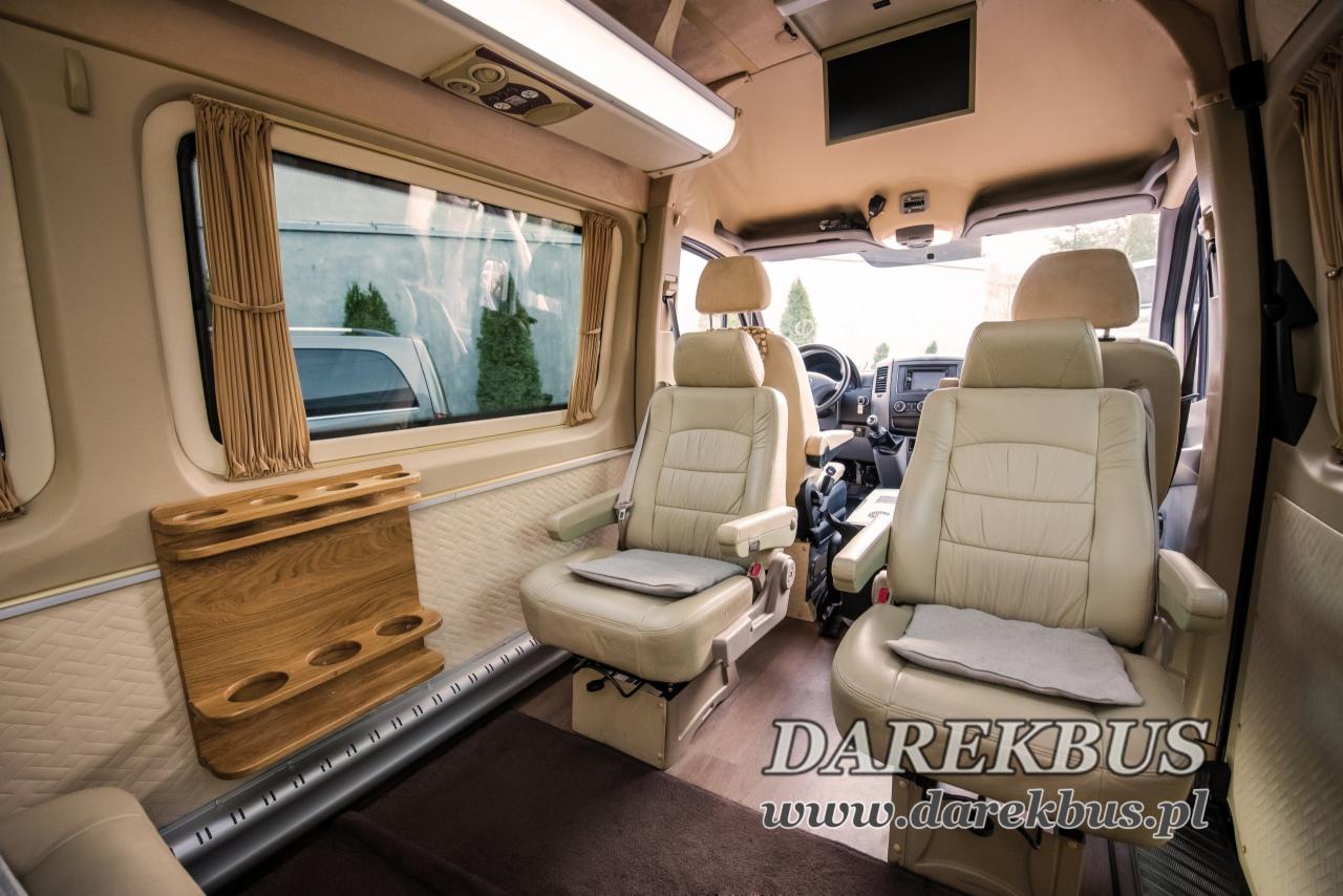 Wnętrze luksusowego busa z beżowymi skórzanymi fotelami, drewnianym uchwytem na napoje przy oknie i telewizorem na suficie.