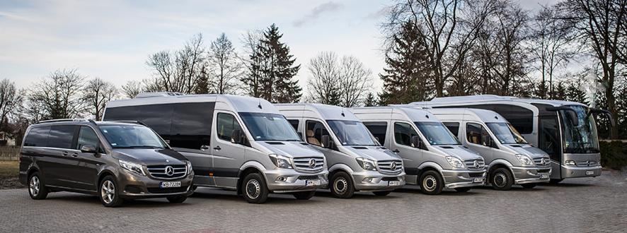 Srebrne i szare busy Mercedes-Benz ustawione w rzędzie na parkingu, widok z przodu, z drzewami w tle.