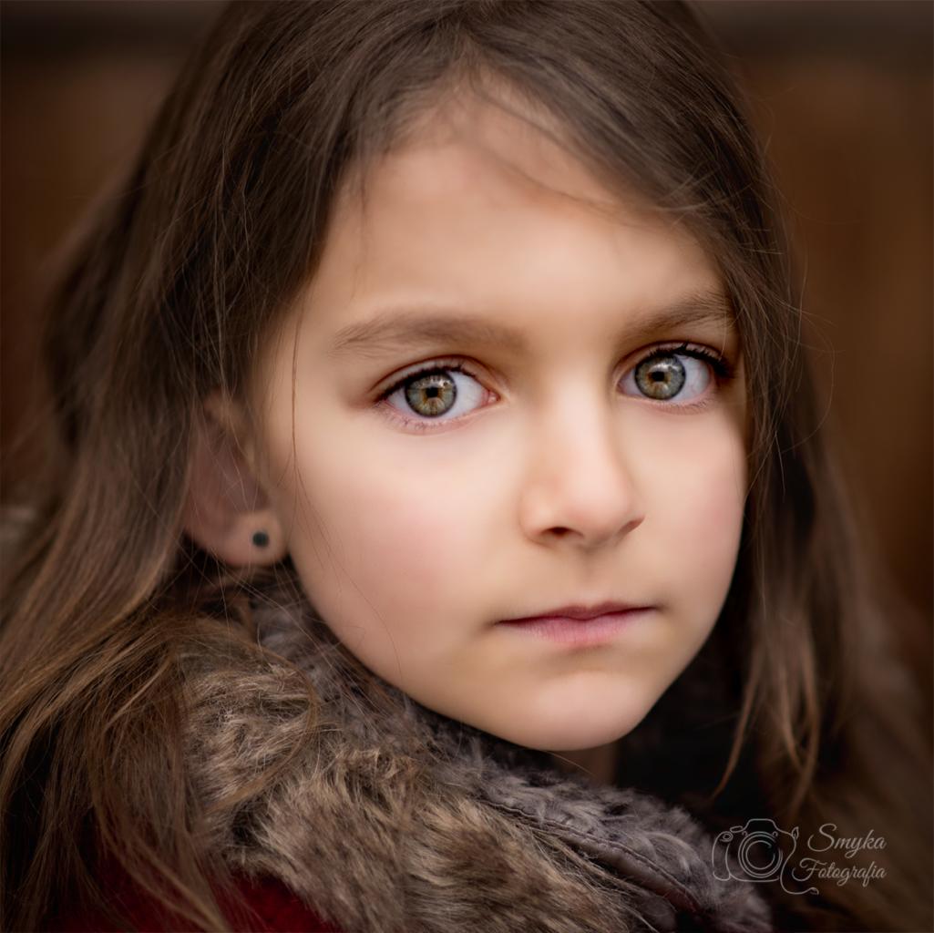 Portret dziewczynki o zielonych oczach i długich brązowych włosach, ubrana w futrzaną kamizelkę, z logo fotografa w prawym dolnym rogu.