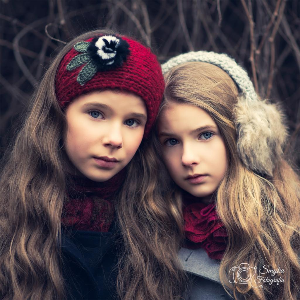 Portret dwóch dziewczynek z długimi włosami i niebieskimi oczami, ubranych w zimowe akcesoria, na tle gałęzi.