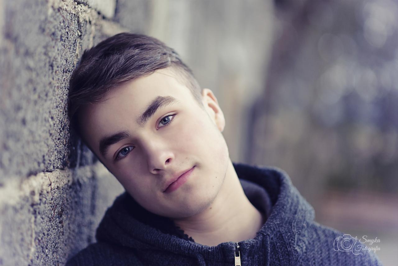 Portret młodego mężczyzny opierającego się o kamienną ścianę w szarej bluzie z kapturem, z subtelnym uśmiechem i jasnymi oczami.