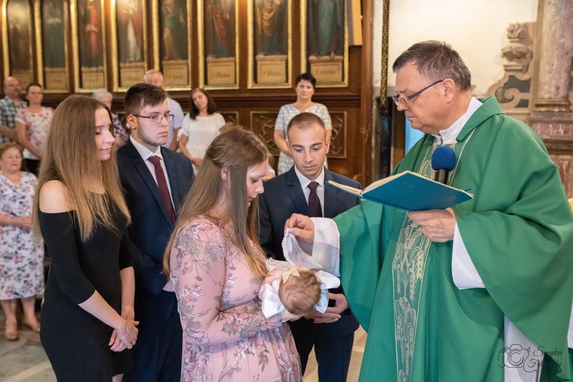 Ksiądz w zielonej szacie polewa wodą główkę niemowlęcia trzymanego przez matkę w sukience w kwiaty, w tle rodzina i obrazy w kościele.