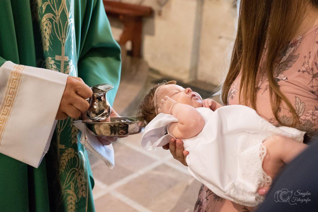 Ksiądz w zielonej szacie trzyma naczynie z wodą, podczas gdy kobieta w kwiecistej sukience trzyma śpiące niemowlę ubrane w białą szatkę.