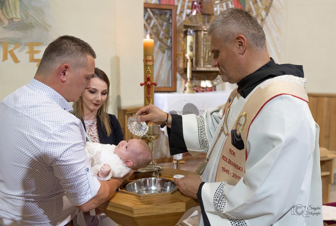 Ksiądz w białej szacie udziela chrztu dziecku trzymanemu przez mężczyznę, w tle kobieta i wnętrze kościoła.