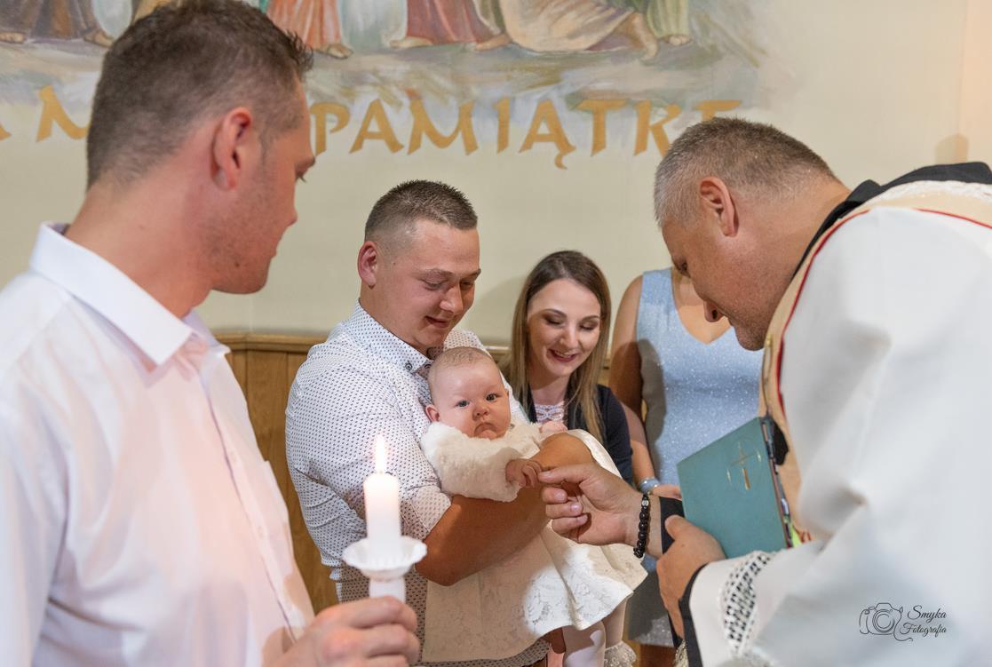 Uśmiechnięci rodzice trzymają dziecko ubrane w białą sukienkę podczas ceremonii chrztu, w tle ksiądz czytający z księgi i inni goście.