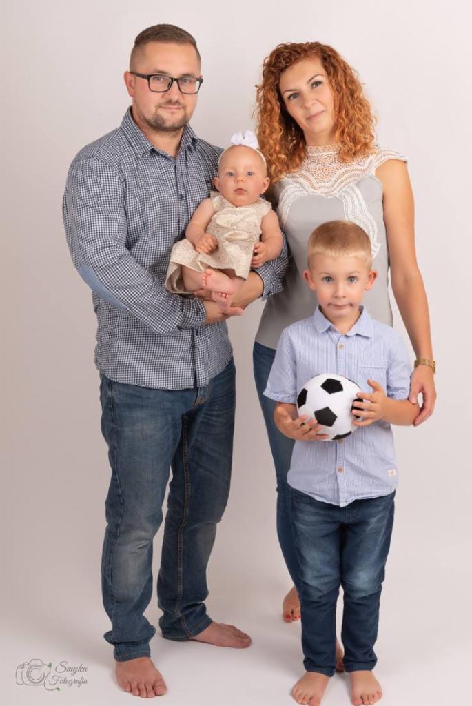 Portret rodzinny: ojciec trzyma niemowlę w beżowej sukience, matka stoi obok syna z piłką, wszyscy boso na jasnym tle.