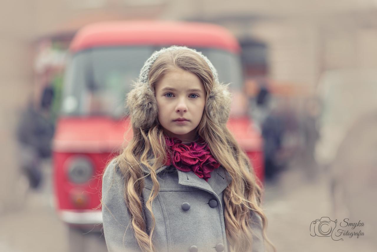 Portret dziewczynki w szarym płaszczu, na tle rozmytego czerwonego busa i innych przechodniów, z futrzanymi nausznikami i bordową falbanką pod szyją, w chłodnej tonacji kolorystycznej.