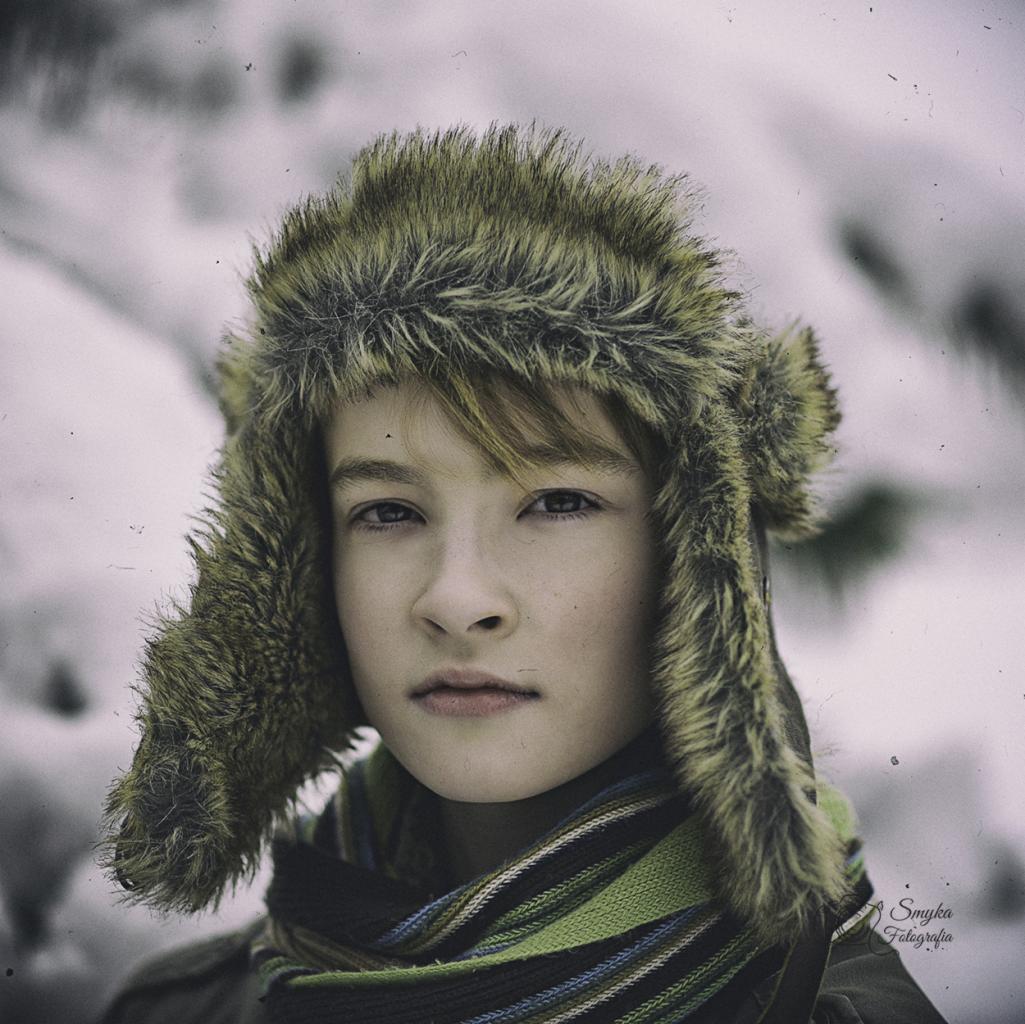 Portret chłopca w futrzanej czapce z nausznikami i pasiastym szaliku na tle ośnieżonego krajobrazu, stylizowany na retro.