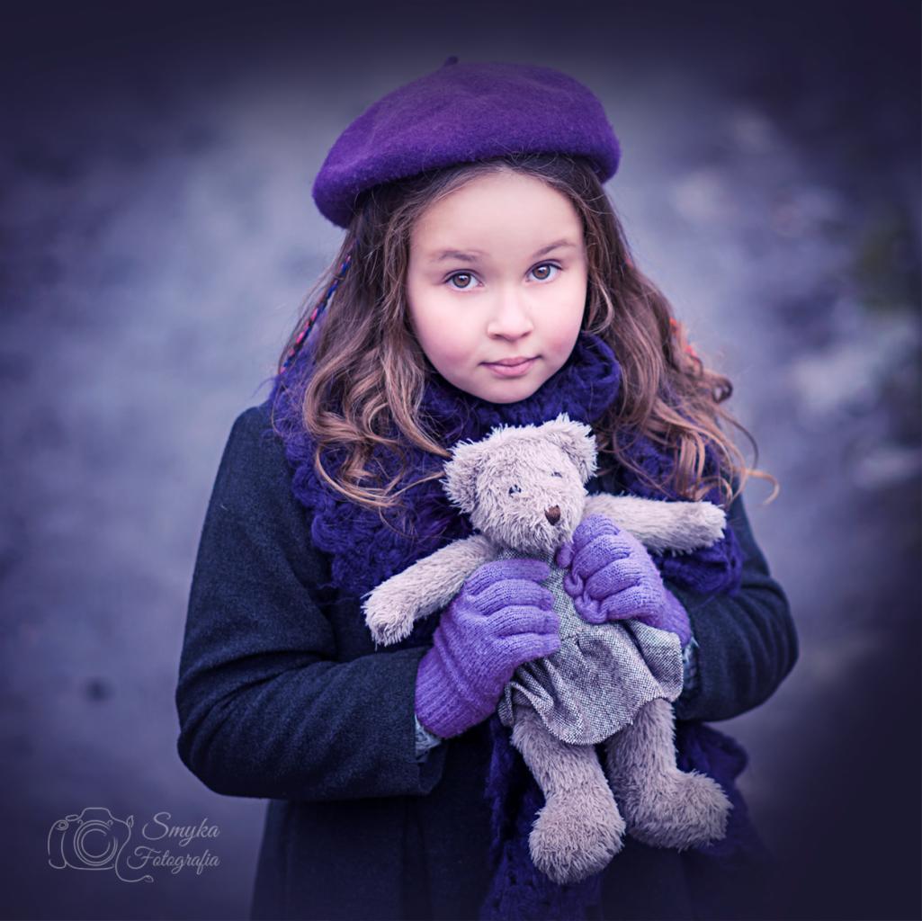 Portret dziewczynki w fioletowym berecie i szaliku, trzymającej pluszowego misia w rękawiczkach, na rozmytym tle.