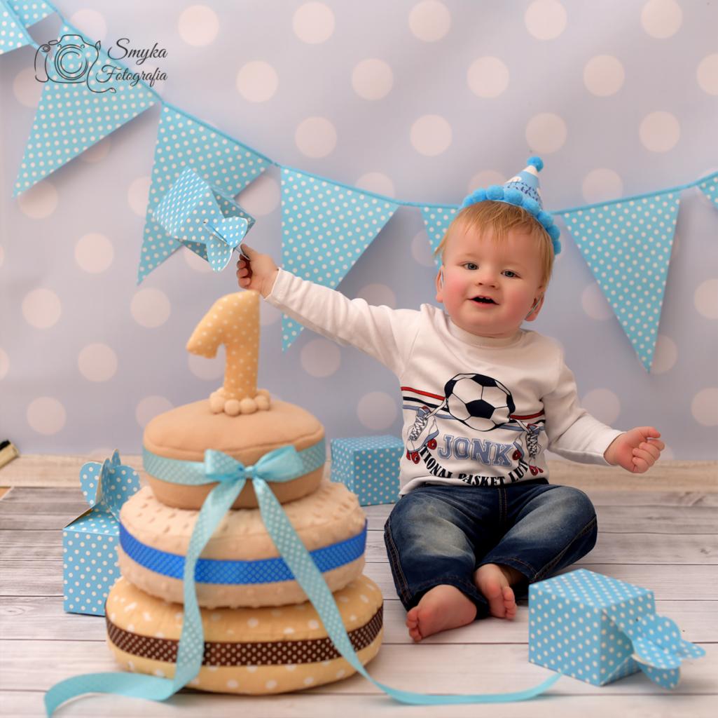 Radosny roczny chłopiec w niebieskim czapeczce, siedzący na podłodze w studio w Krosno, z pluszowym tortem z cyfrą '1' i niebieskimi prezentami w kropki na tle błękitnej girlandy w kropki.