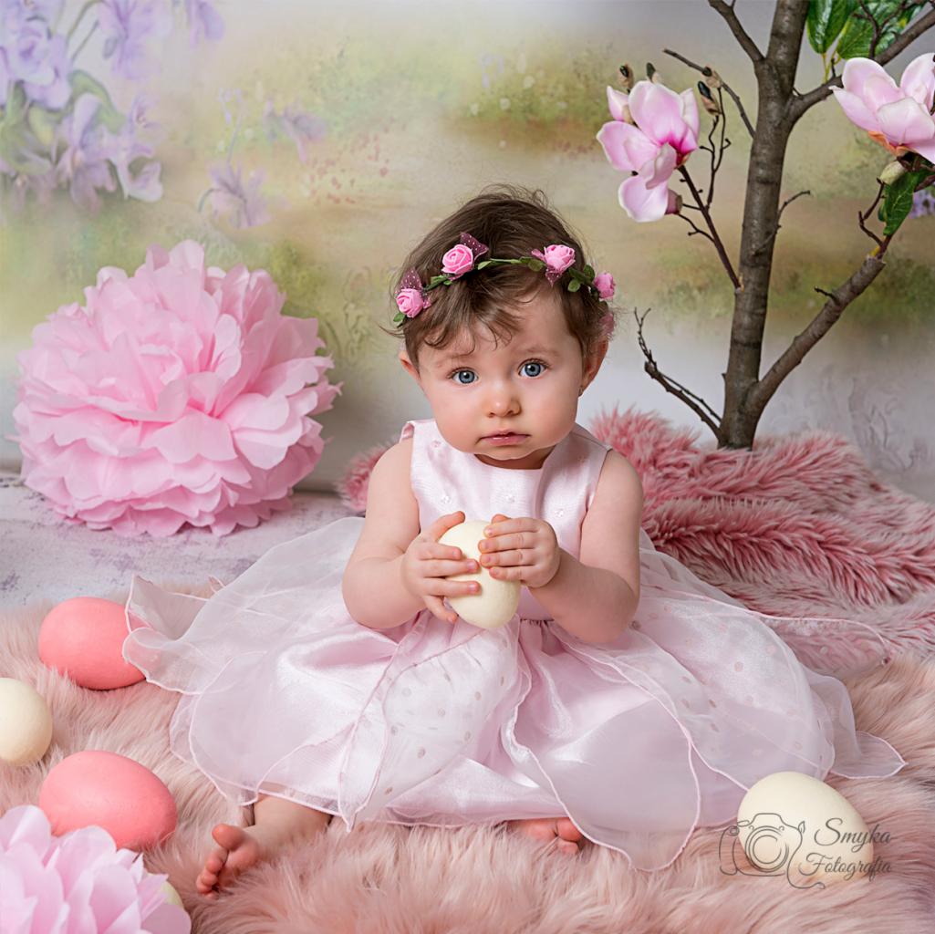 Urocza dziewczynka w różowej sukience i wianku z róż trzyma białe jajko na tle wiosennej dekoracji z kwiatami i pisankami.