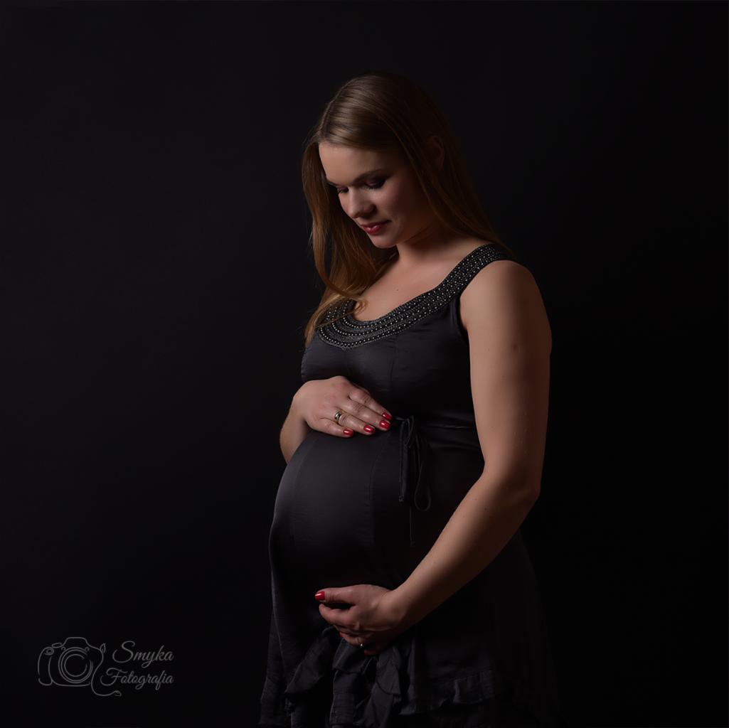 Kobieta w czarnej sukience, z rękami ułożonymi na brzuchu, delikatnie spogląda w dół, ciemne tło studyjne.